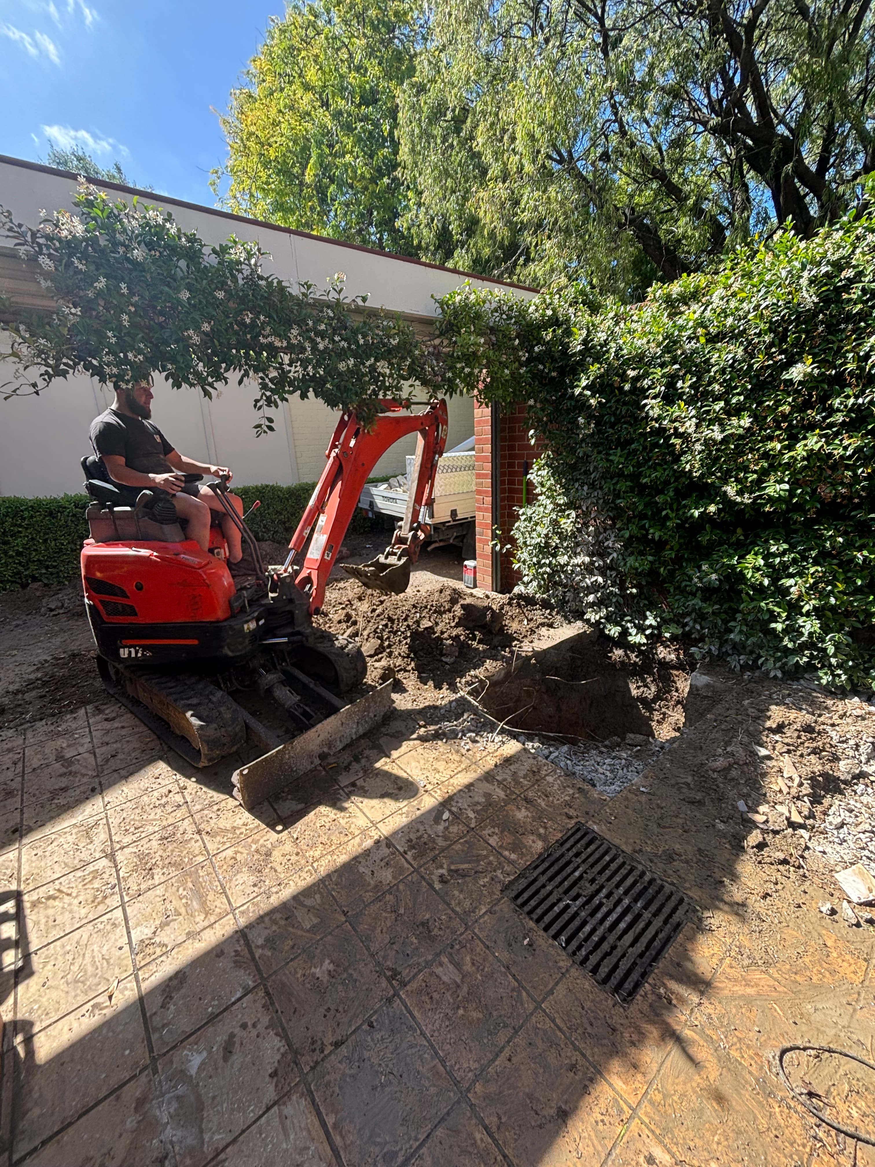 Man using a small red excavator to dig a hole in a paved courtyard.