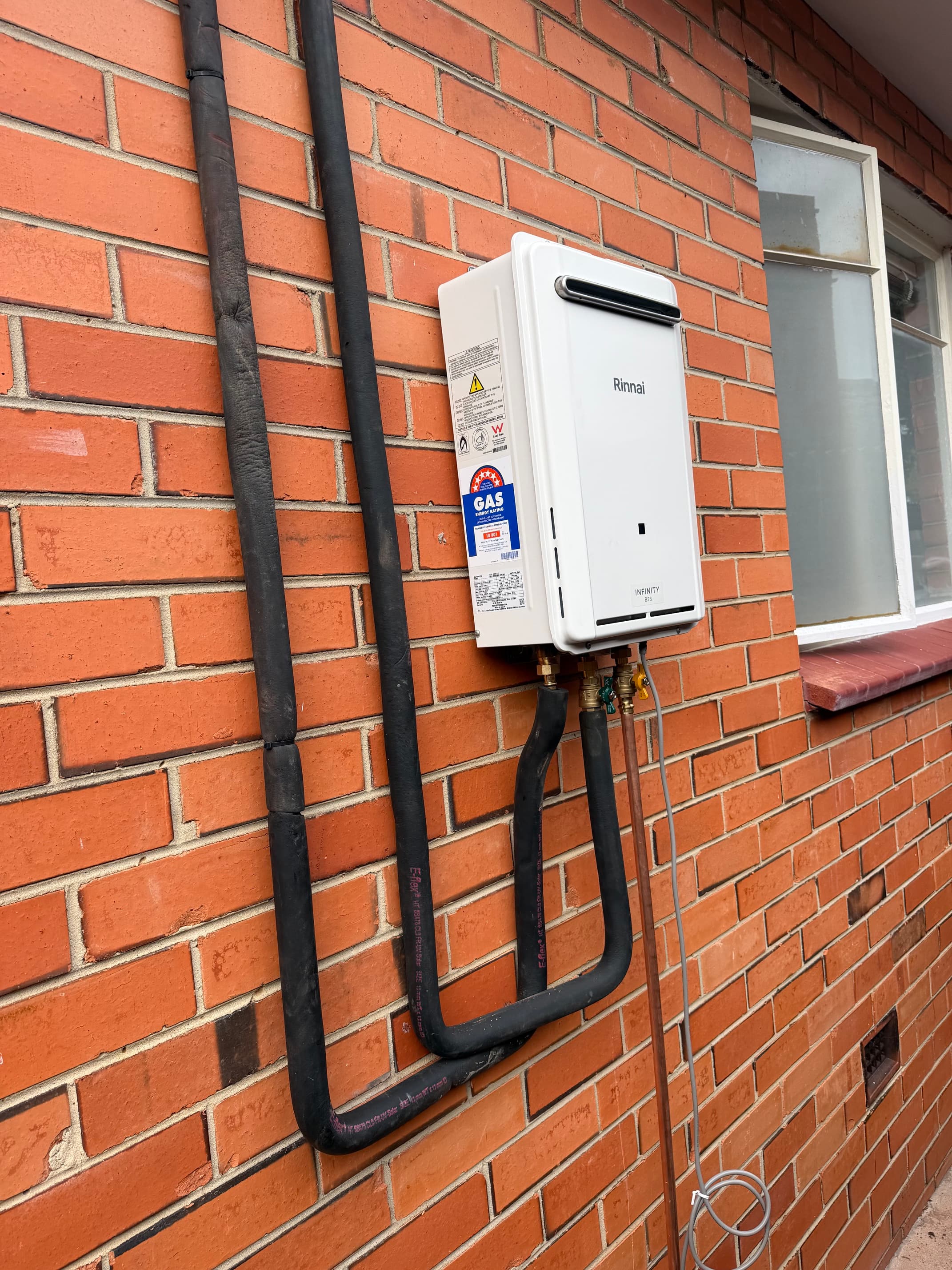 White Rinnai gas hot water system mounted on a red brick wall with black insulated pipes.