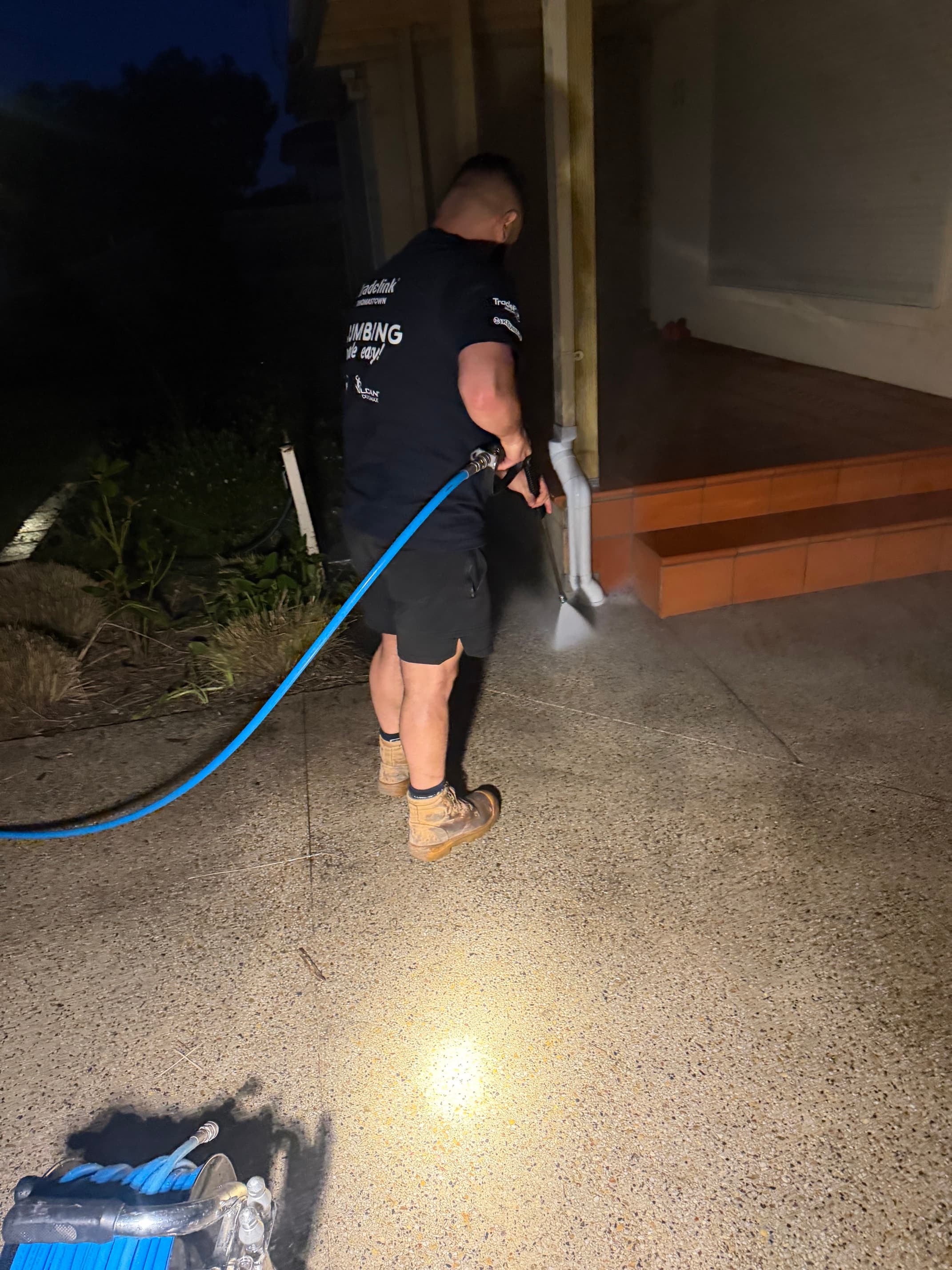 A man in a plumbing shirt pressure washes a concrete driveway at night under bright light.