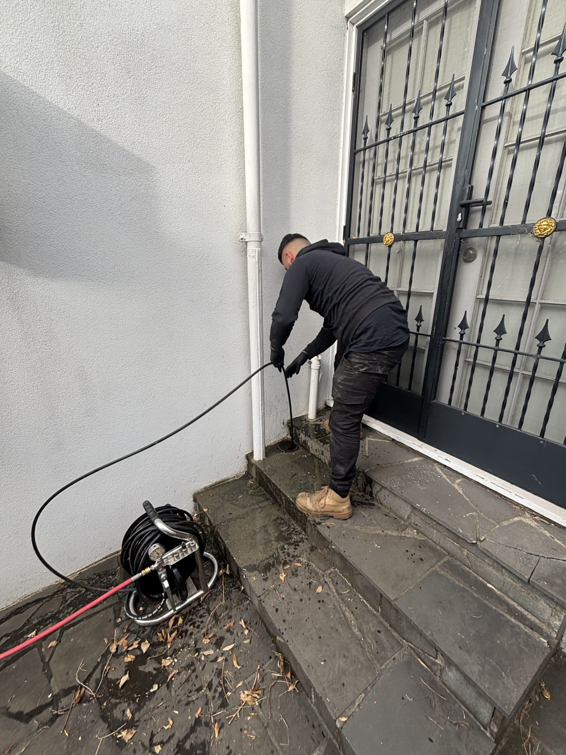 Technician feeds a black hose into a drain on stone steps outside a white building.